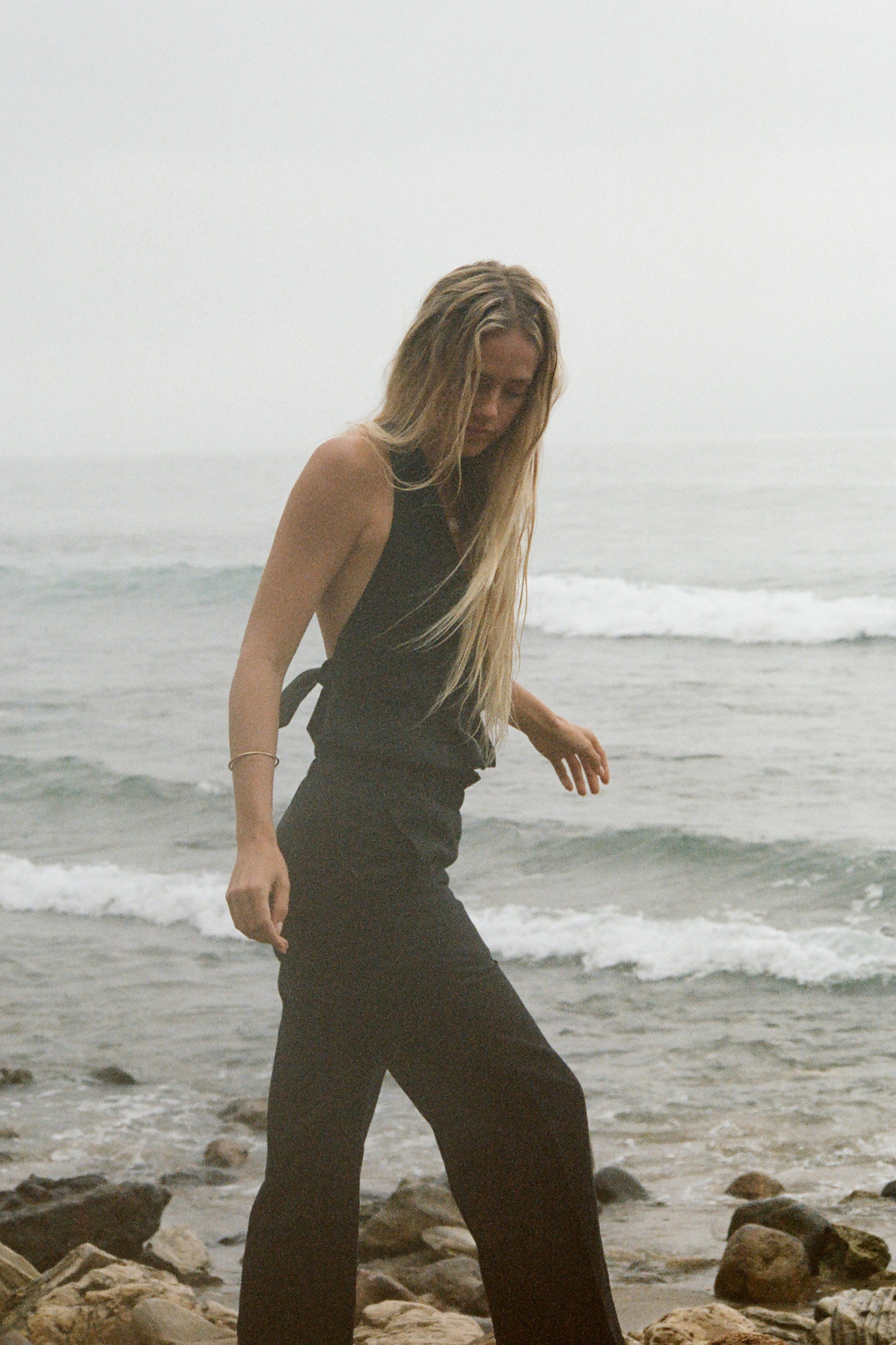 Woman in black set walking along a rocky beach with ocean waves in the background