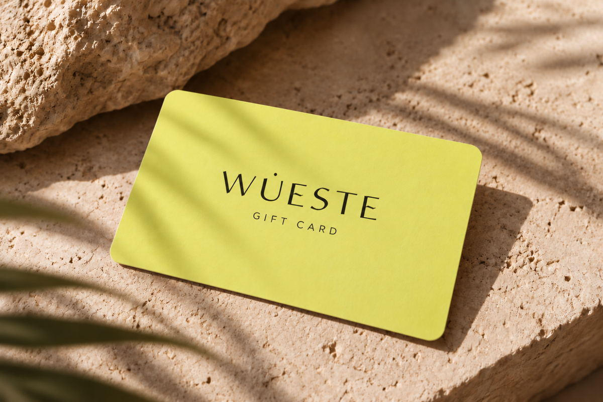 A lime green WUESTE gift card with black text, resting on a textured beige stone surface with natural leaf shadows.